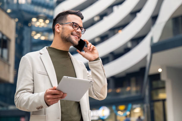 istock Smiling businessman holding digital tablet talking over mobile phone outside office building 2149163878