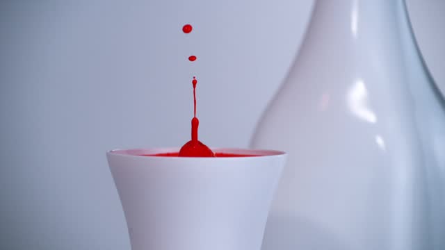 Red drop slowly falls into a white cup. Static view of the quiescent surface response after impact