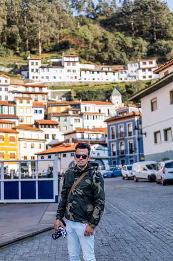 Camouflage hoodie, jeans, and brown bag, Latino tourist exploring Cudillero's vibrant countryside.