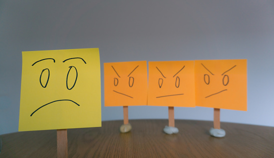 upset and sad faces drawn on sticky notes with gray background, angry faces looking at sad face