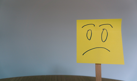 sad face drawn from front on sticky note with gray background, depression