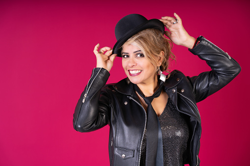 A woman wearing a black jacket and a black hat. She is smiling and holding her hat. The photo has a pink background