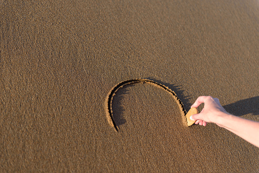 Young woman on the beach is drawing a heart shape on the sand at sunset.