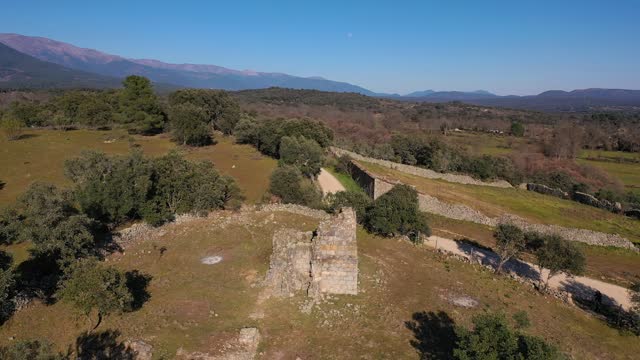 Drone flight in reverse with a medieval ruined hermitage from the 11th century surrounded by fields with pastures and groves, in the background the mountains of the Sierra de Gredos in Avila Spain