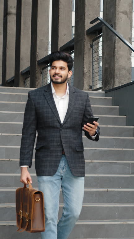 Vertical Screen: Attractive businessman walking outside near big office building and looking around. Muslim businessman in stylish formal wear with brown leather suitcase in hands.