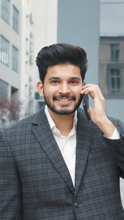 Vertical Screen: Arabian businessman solving urgent working issues on distance. Portrait man. Confident male entrepreneur in stylish formal outfit walking on street with suitcase in hands an talking on mobile phone.