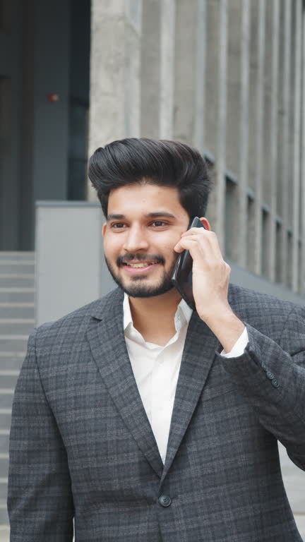 Vertical Screen: Portrait of positive arabian man in formal wear having mobile conversation while standing on city street. Blur background of modern office building. Concept of business