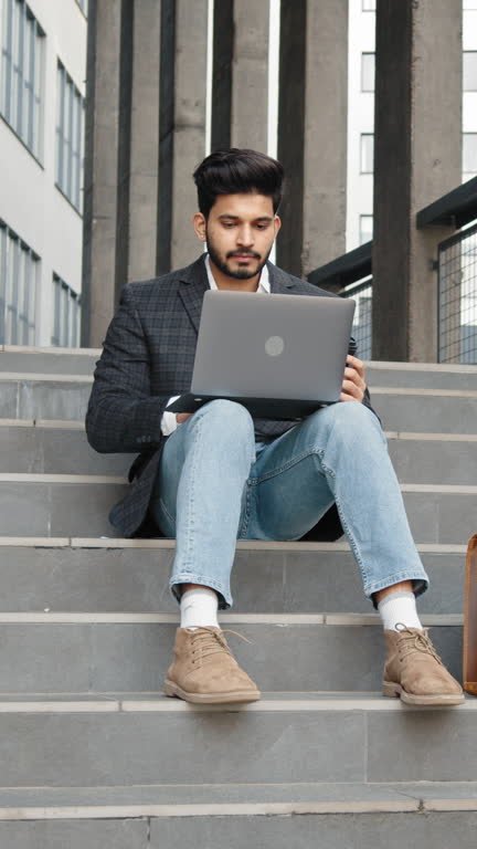 Vertical Screen: Successful muslim man working on laptop on stairs outdoors. Happy arabian freelancer in trendy office clothes showing thumb up while sitting on stairs with wireless laptop on knees.