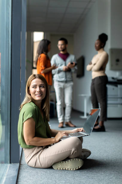 istock Young beautiful female student sitting on the floor and using her laptop computer next to the window 2149176191