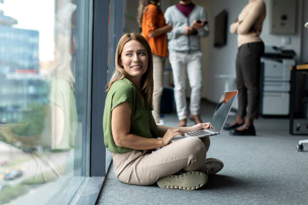 Young beautiful female student sitting on the floor and using her laptop computer next to the window