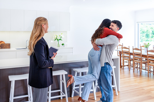 Happy couple embracing and celebrating after buying or renting their new home. The real estate agent is standing beside them with a digital tablet. The kitchen and dining room can be seen in the background