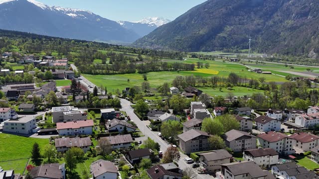 Quaint idyllic landscape of Trimmis Town in Switzerland with homes and buildings in sunlight. Snowy Summits of Alps in background. Aerial forward wide shot.