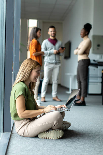 istock Young beautiful female student sitting on the floor and using her laptop computer next to the window 2149176536