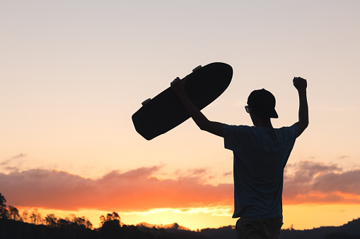 Silhouette of skater man celebrating with his longboard skate, with sunset sky in the background. Conveys the feeling of freedom and well-being, promoting a healthier and happier lifestyle through outdoor activities.