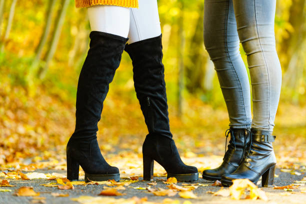 Close-up view of a person's black leather boots with block heels. The boots are positioned on a stone wall, surrounded by a ground cover of brown and orange autumn leaves.