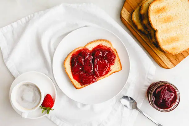 Slice of toasted white bread and homemade berry preserves with coffee on white table