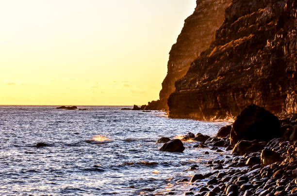 The ocean is calm and the sky is orange. The rocks on the shore are large and the water is clear