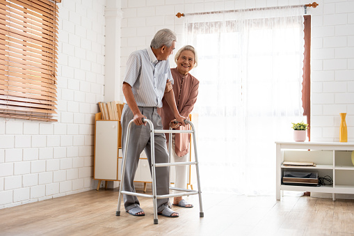 Asian attractive senior woman support elderly man walking with walker. Happy family, Beautiful mature older couple doing physical therapy after retirement for health together in living room at home.