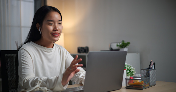 Selective focus, Young Asian woman wear wireless headphones sitting at desk video call with a colleague on a laptop in home office, working overtime, working from home at night