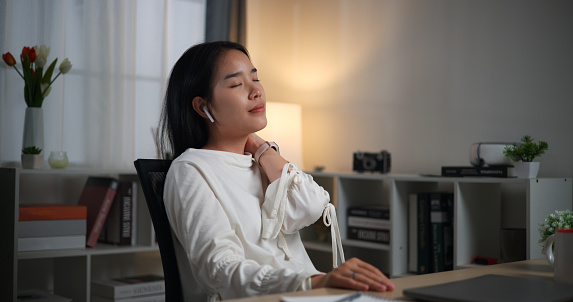 Selective focus, Young Asian woman wear wireless headphones working late night and stretches to relieve fatigue while sitting at desk in home office, working overtime, working from home at night