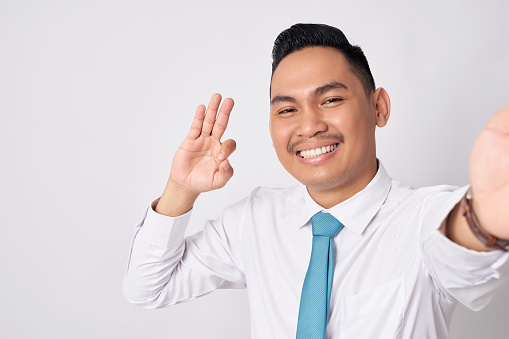 Close up smiling happy fun young Asian businessman wearing a formal shirt tie and showing okay sign isolated on white background. Taking a selfie shot on the phone