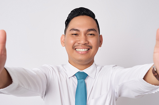 Close up smiling happy fun young Asian businessman wearing a formal shirt and tie isolated on white background. Taking a selfie shot on the phone