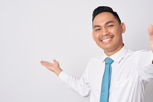 Close up smiling happy fun young Asian businessman wearing a formal shirt tie and showing empty space with hand isolated on white background. Taking a selfie shot on the phone