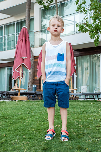 Sad boy in beach clothes stands on a well-maintained lawn on background of modern seaside hotel with closed red umbrellas and lounge chairs