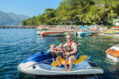 Father with two small sons wearing life jackets is seated on a jet ski near beach with boats and pine trees in a seaside resort