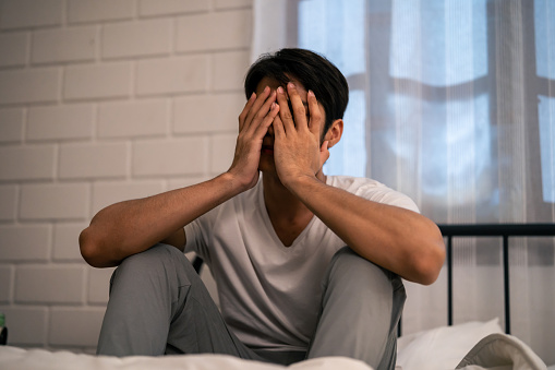 Asian frustrated insomnia man in pajamas sit on bed in dark night time. Attractive depressed young sleepless man feeling upset, worried and concerned cannot sleep from nightmare in bedroom in home.