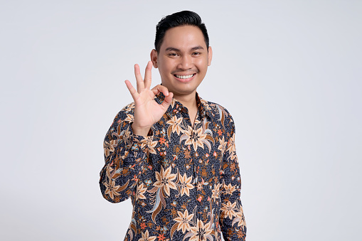 Smiling happy young handsome Asian man wearing a batik shirt doing ok sign with fingers isolated on white background