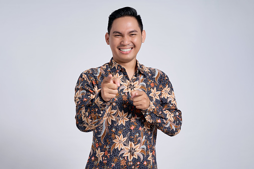 Portrait of smiling young asian man wearing batik shirt looking at the camera and pointing index finger at you isolated on white background