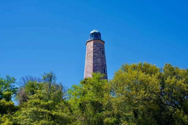 Fort Story, Virginia, USA - April 13, 2024: Trees in the foreground partially obscure the Old Cape Henry Lighthouse on a sunny day.