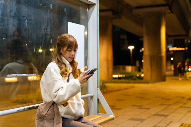 Asian woman using smartphone with mobile app text message during waiting for bus at bus stop. Attractive girl enjoy urban outdoor lifestyle travel city street with wireless technology device.