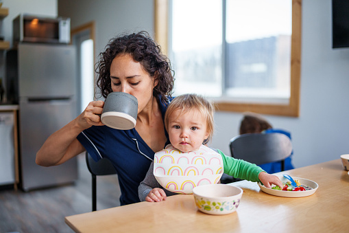 An adorable one year old Eurasian toddler boy looks directly at the camera while sitting on his mother's lap and eating breakfast. The sleepy looking woman is wearing pajamas and taking a sip of coffee.