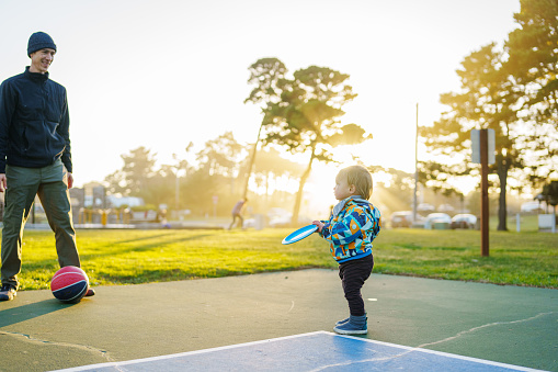 A cute one year old Eurasian boy plays with a disc golf frisbee as his dad shoots some baskets at an outdoor basketball court at a park in Oregon on a sunny and cool afternoon.