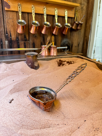 Copper cezve with fresh Turkish coffee on sandy stove, traditional copper coffee pots on wooden rack in background, symbolizing the authenticity of Middle Eastern coffee traditions. High quality photo