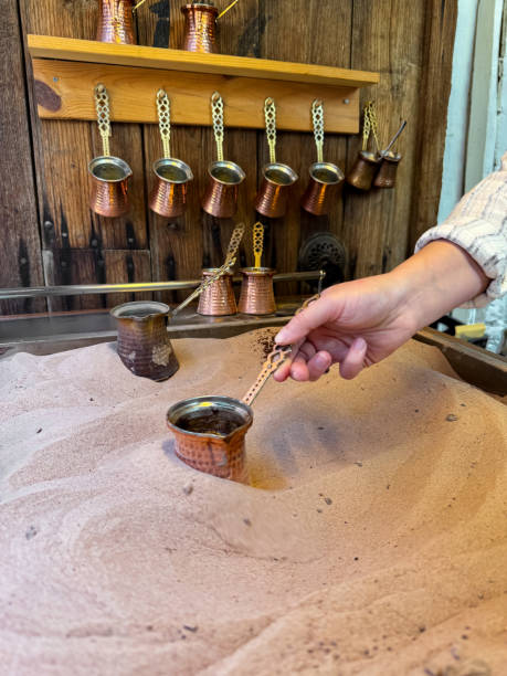 istock Preparing Turkish coffee in traditional copper pot on sandy stove, with hand holding the handle, copper cezves and wooden wall rack in the background. Turkish coffee culture concept. 2149207146