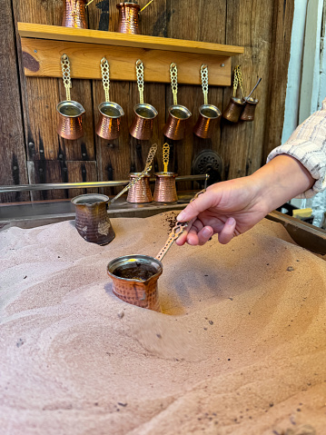 Preparing Turkish coffee in traditional copper pot on sandy stove, with hand holding the handle, copper cezves and wooden wall rack in the background. Turkish coffee culture concept. High quality