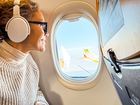 Smiling passenger with Bluetooth noise-canceling headphones looking out of the airplane's porthole using the on-board Wi-Fi Internet connection. Middle-aged woman sitting by the window relaxing while waiting to arrive