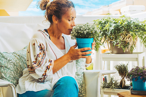 Beautiful lady takes care of her plants on the balcony. Sitting blonde lady admires her flowers. Hobby and botanical care concept