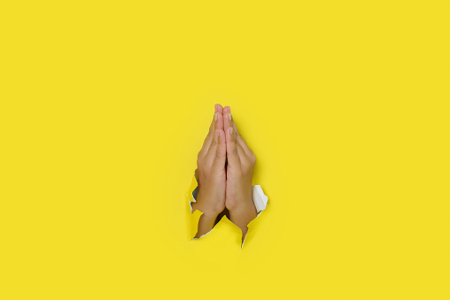 Female hands in prayer gesture coming out of a hole in yellow paper. Copy space.