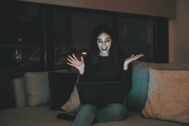 A beautiful young woman using laptop for video calls with a bright screen late at night at home. Smiling girl smiles and gestures while talking to family and friends.