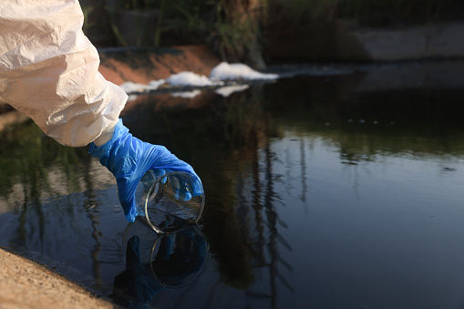 Close-up scientist hand with a white PPE suit and nitrile gloves collect sample waste water into a flask for laboratory water analysis, industrial and environmental concept