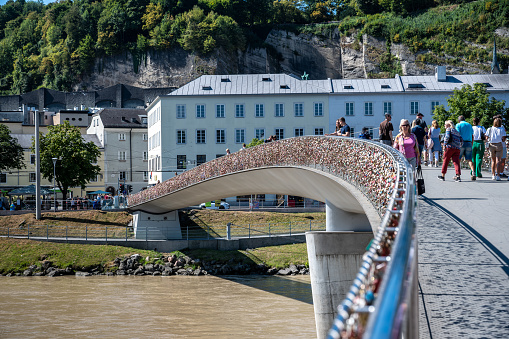 Salzburg, Austria, August 15, 2022. The pedestrian bridge of love with padlocks glistening under the summer sun. People cross it, a couple stopped to observe the view.