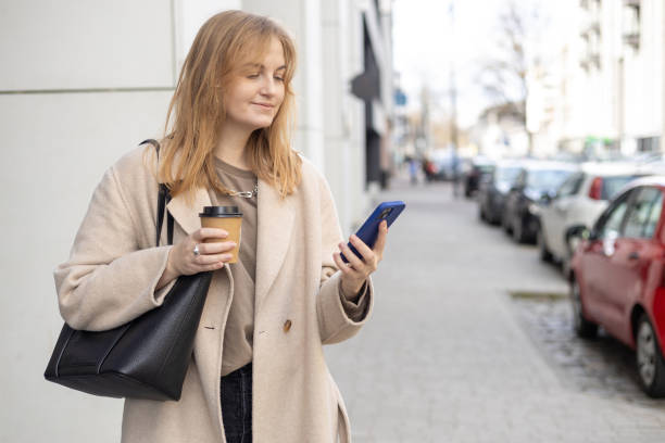istock Beautiful smiling female with coffee cup using smartphone and standing outdoors. Phone Communication. Happy cheerful young woman walking on city street, Urban lifestyle concept. Traveler 2149211302