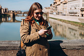Happy 30s girl using smartphone on famous Old bridge in Florence. Stylish woman visiting Italian landmarks. Concept of travel, tourism and vacation in city. Woman walking on city street checks phone.