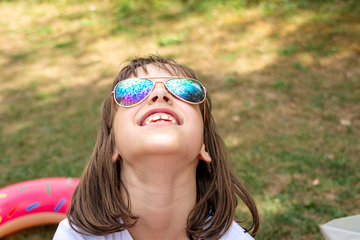 Little girl wearing sunglasses and looking up the sky while sitting at riverbank during summer day
