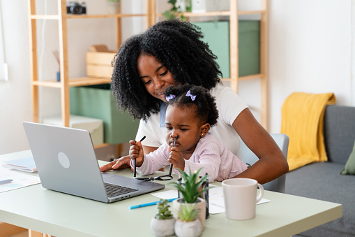 African mother assists her young daughter at a laptop in their cozy home workspace