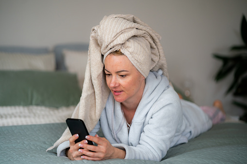 Woman in bathrobe and towel on her head lying on bed and using mobile phone at home